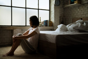 A depressed woman sits on the floor in a bedroom with her knees pulled to her chest