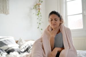 A woman sits on bed and rubs her dry throat